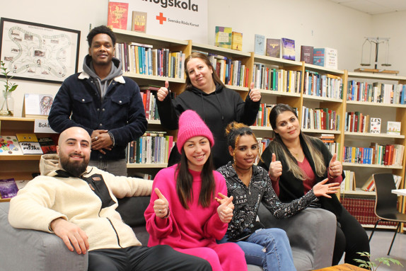 En grupp glada deltagare på Röda Korsets folkhögskola poserar för kameran i skolans bibliotek. Några sitter i en grå soffa andra står bakom. De säger välkomna till folkhögskolan!