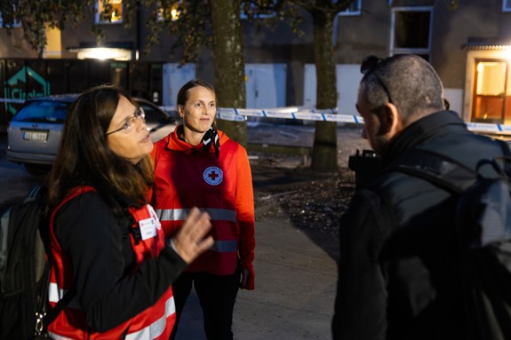 Tre kvinnor i Röda Korsvästar står på torget i Skärholmen. En av kvinnorna omfamnar en kvinna som söker stöd. En annan av Röda Korspersonerna hälsar på en man.